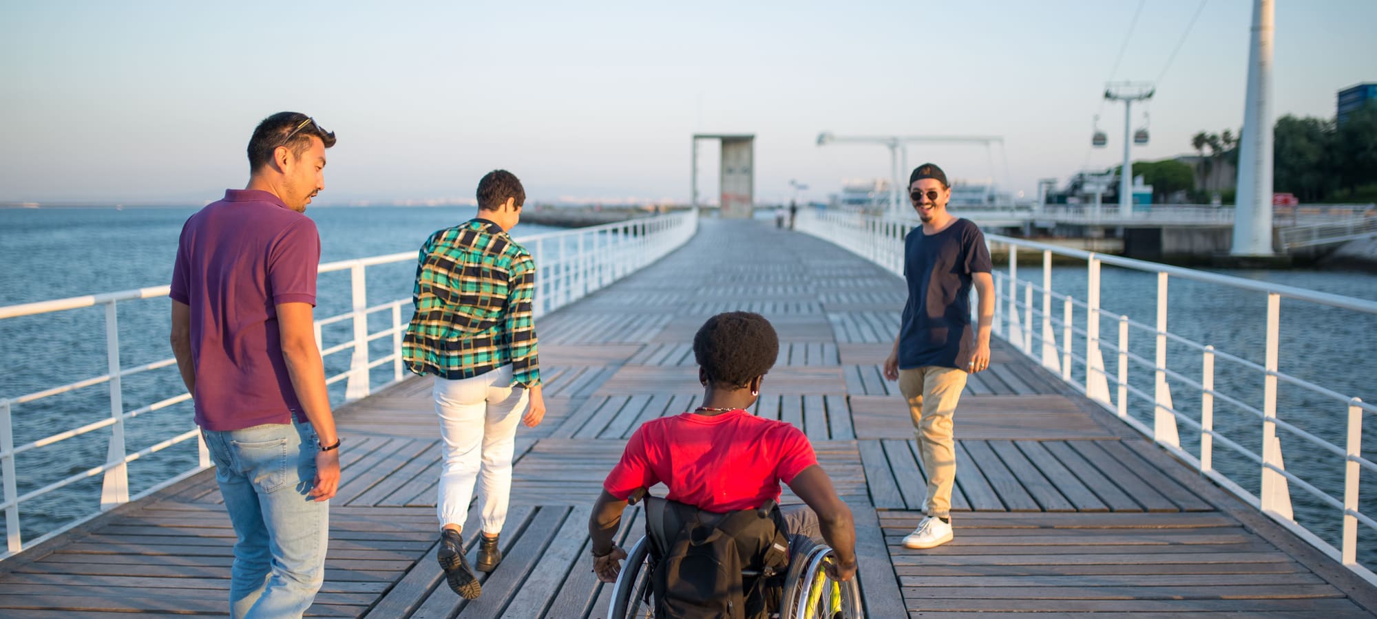 Friends walking along a harbour boardwalk in the sun, one of them using a wheelchair