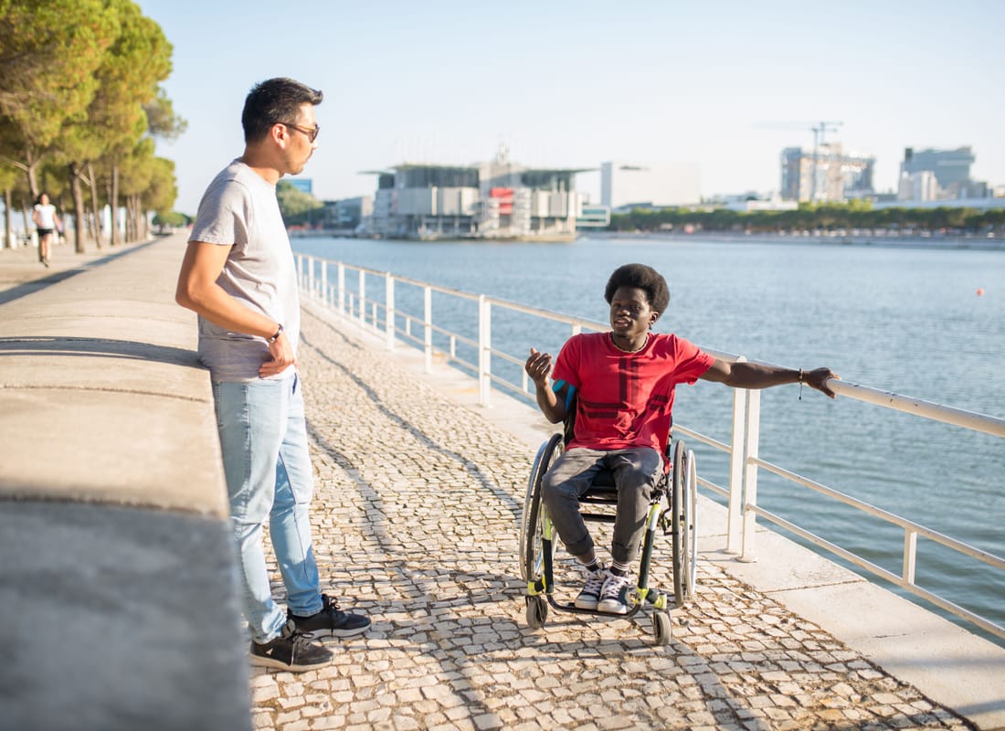 Two men talking beside the water, one using a wheelchair