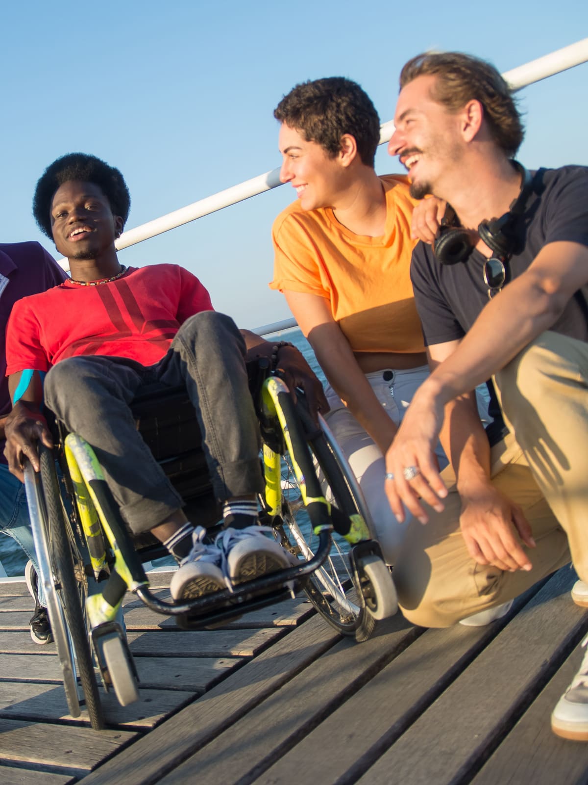 Four friends laughing together on a seaside boardwalk, one of them using a wheelchair